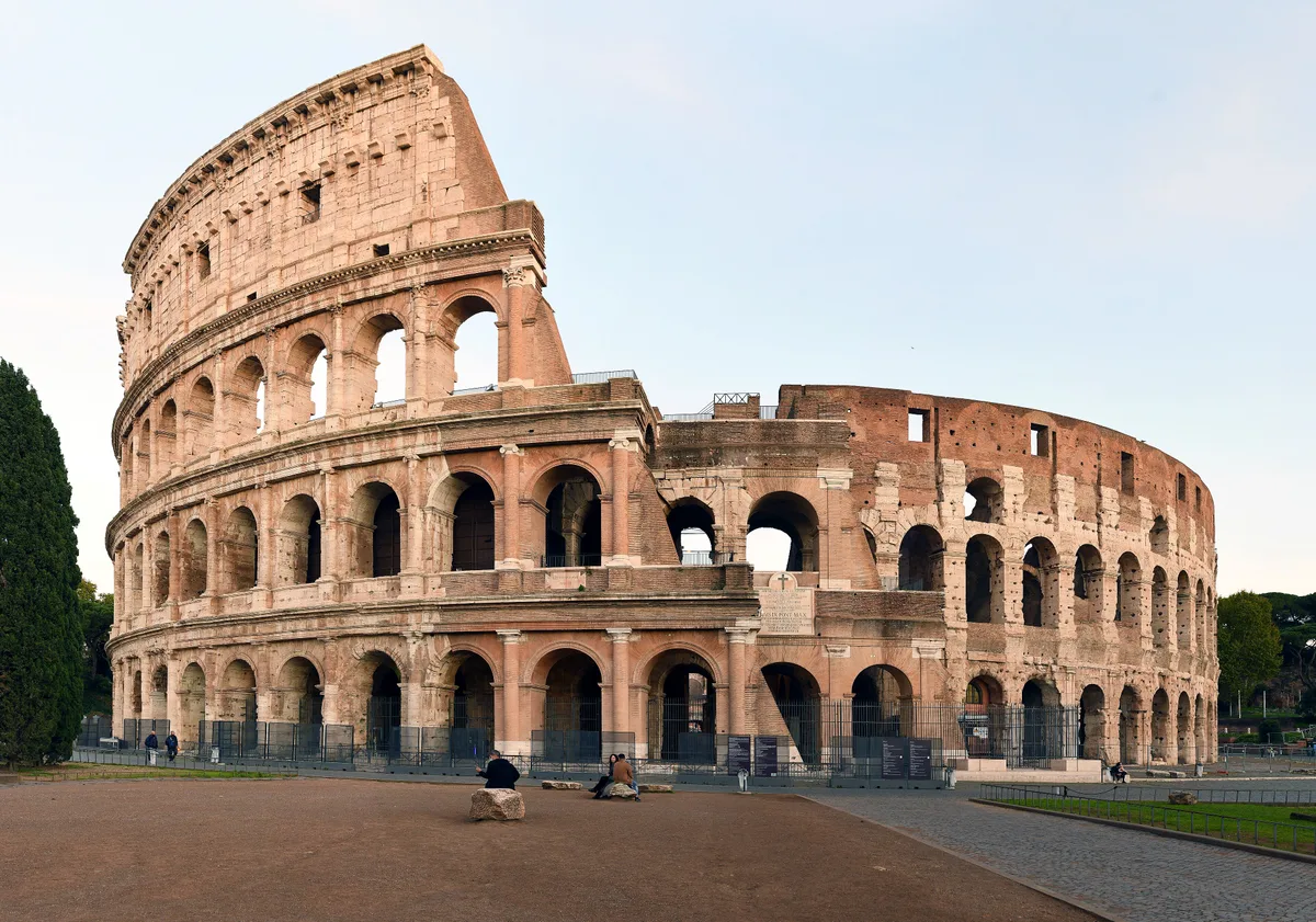 The Colosseum in Rome - An elliptical amphitheatre of travertine stone rising four stories, with rows of arched...